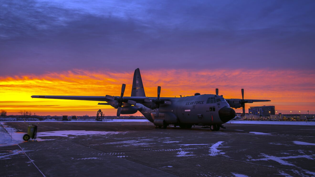 U.S. Air Force C-130 Hercules on a snowy runway at sunset with a vibrant orange and purple sky.