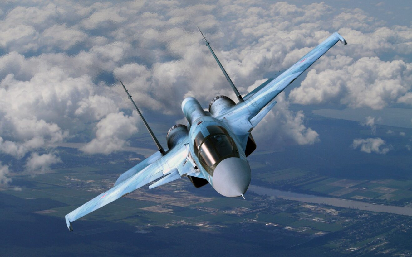 A twin-engine fighter jet, likely a Sukhoi, flies above the clouds with farmland visible below, showcasing its sleek design and blue camouflage.
