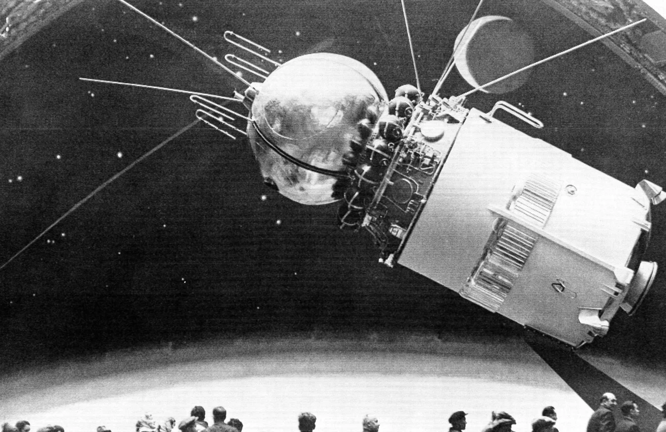 Black and white image of a Soviet Luna spacecraft model displayed in front of an audience, illustrating early lunar exploration technology with detailed antennas and equipment.