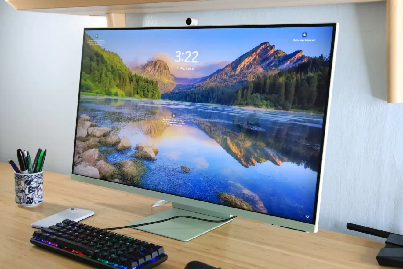 Samsung Smart Monitor M8 on a wooden desk, displaying a scenic mountain and lake wallpaper, with a mechanical RGB keyboard, smartphone, and pen holder nearby.
