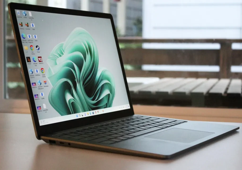 A sleek Microsoft Surface Laptop 5 placed on a white table near a window, displaying the Windows 11 home screen with several app icons on the left side.