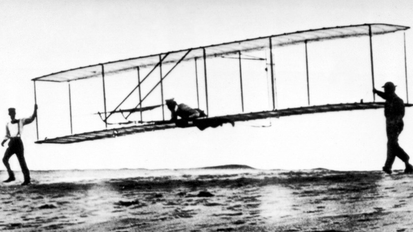 Historic black and white photograph capturing one of the Wright brothers piloting the Wright Flyer during its first powered flight in 1903 at Kitty Hawk, North Carolina. The pilot lies face down on the lower wing structure of the biplane, suspended above sandy ground with the aircraft in midair. This iconic moment marks the birth of modern aviation.