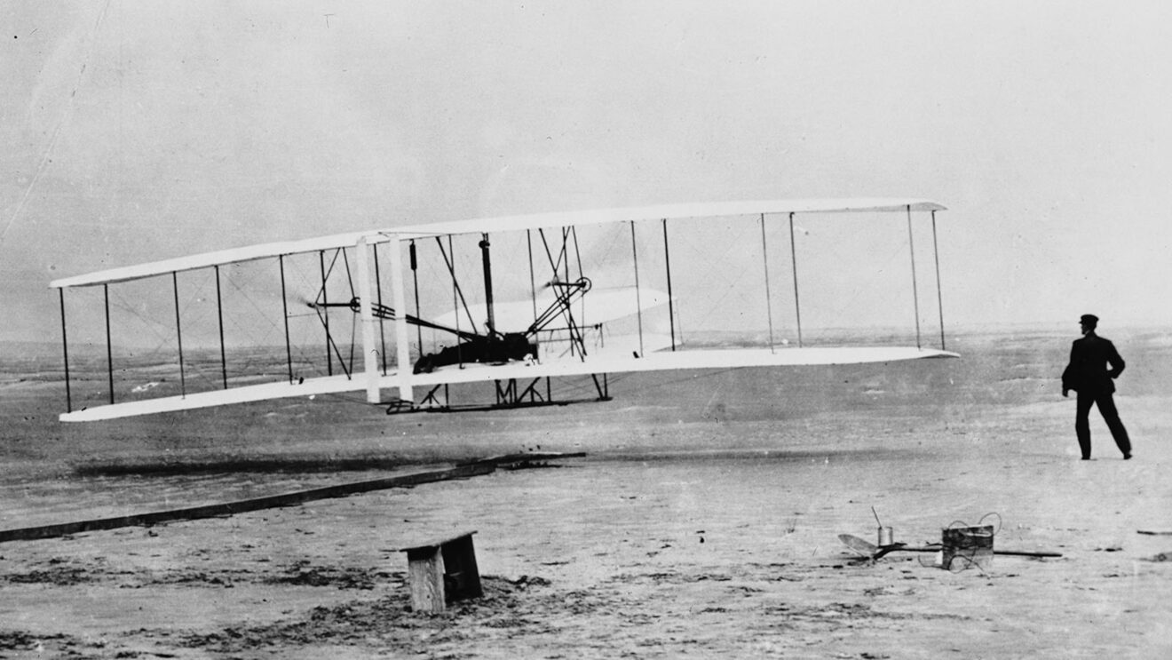Historic photograph of the Wright brothers' first powered flight on December 17, 1903, at Kitty Hawk, North Carolina. The Wright Flyer, a biplane with a wingspan supported by struts and wires, is seen lifting off the ground with one brother piloting the aircraft while the other watches from the sandy terrain. This iconic image captures the groundbreaking moment that marked the dawn of modern aviation.